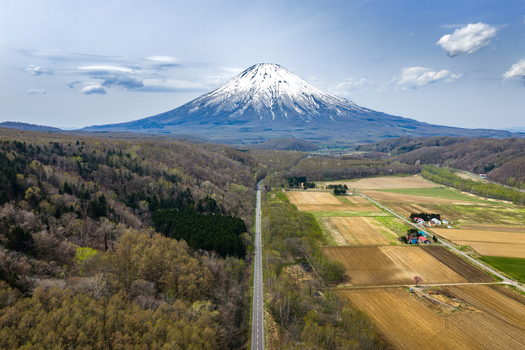 Aerial view of Mount Yotei with its snow capped peak rising above the surrounding forests agricultural fields and a straight road leading towards the horizon in Niseko Hokkaido Japan.