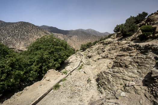 View of a narrow irrigation canal winding through the rocky mountain slopes and lush green trees of the Atlas Mountains in Tizi Oussem Marrakech Safi Morocco. View of a narrow irrigation canal winding through the rocky mountain slopes and lush green trees of the Atlas Mountains in Tizi Oussem Marrakech Safi Morocco.
