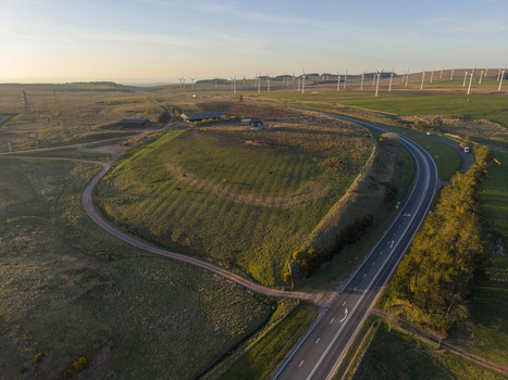 Aerial view of a winding road with a truck passing through green fields and farm buildings with a large wind farm on the horizon under a clear sky. Aerial view of a winding road with a truck passing through green fields and farm buildings with a large wind farm on the horizon under a clear sky.