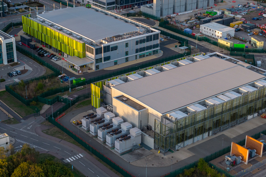 Aerial view of the Kao Data center featuring modern industrial buildings with green facade panels cooling units and a fenced perimeter in Harlow England United Kingdom. Aerial view of the Kao Data center featuring modern industrial buildings with green facade panels cooling units and a fenced perimeter in Harlow England United Kingdom.