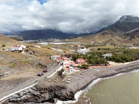 Aerial view of a coastal road winding past white walled buildings with red tiled roofs meeting the dark rocky shoreline under a sky of swirling clouds Puerto de la Aldea Canarias Spain. Aerial view of a coastal road winding past white walled buildings with red tiled roofs meeting the dark rocky shoreline under a sky of swirling clouds Puerto de la Aldea Canarias Spain.