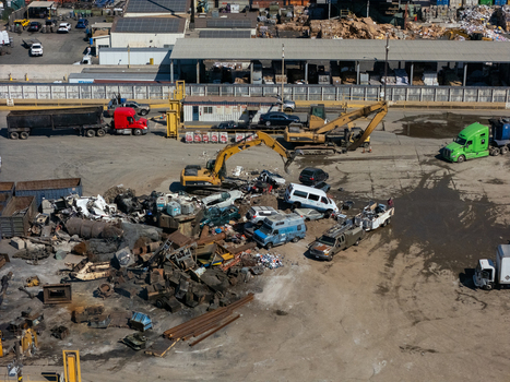 Aerial view of a scrap metal recycling yard featuring excavators sorting through piles of crushed vehicles and industrial waste in Carson California United States.
