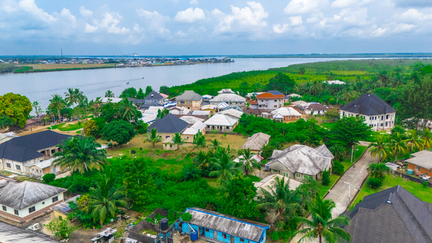 Aerial view of a coastal village with houses featuring dark roofs surrounded by lush tropical vegetation and palm trees along a river in Minima Rivers Nigeria.
