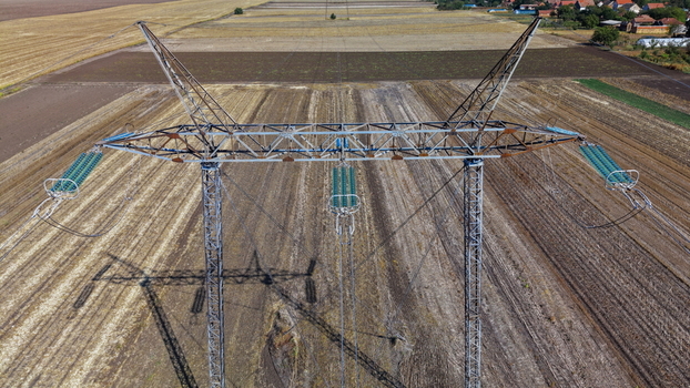 Aerial view of electric power transmission lines standing in a vast agricultural field with brown soil and distant rural houses in Pancevo Serbia.