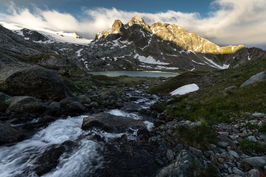 View of a rushing mountain stream flowing towards a glacial lake beneath rugged snow dusted peaks and a cloudy sky in the Alps Aosta Valley Italy. View of a rushing mountain stream flowing towards a glacial lake beneath rugged snow dusted peaks and a cloudy sky in the Alps Aosta Valley Italy.