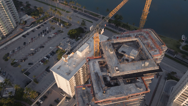 Aerial view of a construction site with a tall yellow crane towering over a high rise building under development adjacent to a parking lot and waterfront in Miami Beach Florida United States. Aerial view of a construction site with a tall yellow crane towering over a high rise building under development adjacent to a parking lot and waterfront in Miami Beach Florida United States.