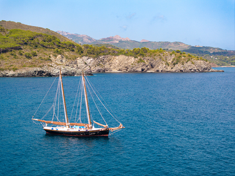 Aerial view of a classic sailboat sailing on the deep blue sea near the rocky coastline and green hills of Lacona. Lacona Tuscany Italy.