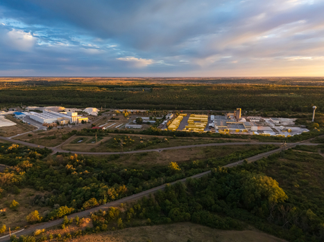 Aerial view of an industrial logistics facility with stacked yellow containers rail lines woodland and farmland beneath a cloudy summer sunset in Visonta Hungary.