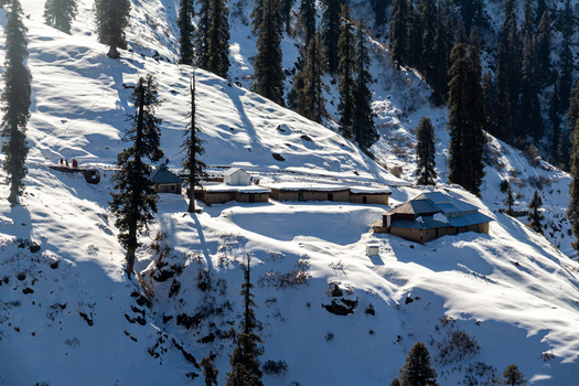 View of snow blanketed mountain slopes dotted with buildings and tall dark trees under a bright sky creating a serene winter landscape Lasdana Azad Kashmir Pakistan.