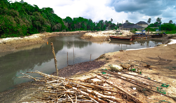 Aerial view of a riverbank in Buguma with wooden boats moored in the muddy water surrounded by lush vegetation and scattered debris in Buguma Rivers Nigeria.