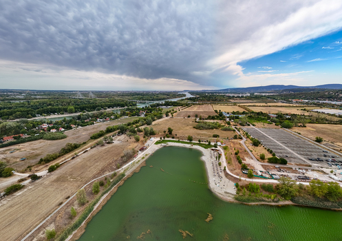 Aerial view of Lake Lupa s green water and sandy beach beside the Danube with Megyeri Bridge farmland a parking area and suburban development in Budakalasz Hungary. Aerial view of Lake Lupa s green water and sandy beach beside the Danube with Megyeri Bridge farmland a parking area and suburban development in Budakalasz Hungary.