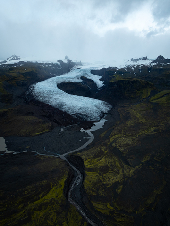Aerial view of Heinabergsjokull glacier with its icy tongue flowing through dark volcanic terrain and mossy green landscapes in Sveitarfelagid Hornafjordur Iceland.