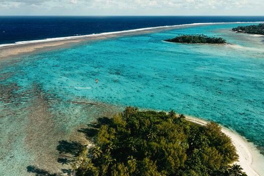 Aerial view of the turquoise lagoon and coral reef surrounding Rarotonga with a kite surfer gliding over the clear water and lush tropical vegetation on the island in Cook Islands. Aerial view of the turquoise lagoon and coral reef surrounding Rarotonga with a kite surfer gliding over the clear water and lush tropical vegetation on the island in Cook Islands.