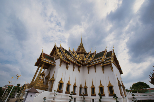 View of The Grand Palace featuring ornate gold trimmed architecture and white walls under a cloudy sky in Bangkok Thailand. View of The Grand Palace featuring ornate gold trimmed architecture and white walls under a cloudy sky in Bangkok Thailand.