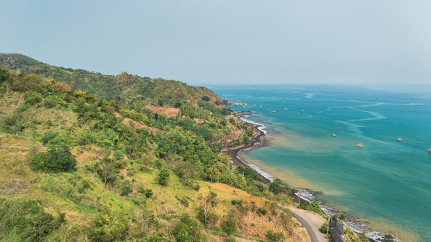 Aerial view of the lush green hills meeting the dark sand beach and turquoise waters of the coast with small boats floating in the sea near Sukabumi Indonesia. Aerial view of the lush green hills meeting the dark sand beach and turquoise waters of the coast with small boats floating in the sea near Sukabumi Indonesia.