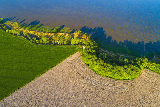 Aerial view of the coastline in Vanersnas featuring green fields tilled land and trees bordering the lake under warm sunlight Vanersnas Vastra Gotalands lan Sweden. Aerial view of the coastline in Vanersnas featuring green fields tilled land and trees bordering the lake under warm sunlight Vanersnas Vastra Gotalands lan Sweden.