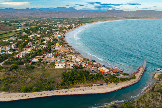 Aerial view of the coastal town of Punta Perula featuring a long pier extending into the turquoise waters of the bay surrounded by buildings palm trees and rolling hills in Punta Perula Jalisco Mexico. Aerial view of the coastal town of Punta Perula featuring a long pier extending into the turquoise waters of the bay surrounded by buildings palm trees and rolling hills in Punta Perula Jalisco Mexico.