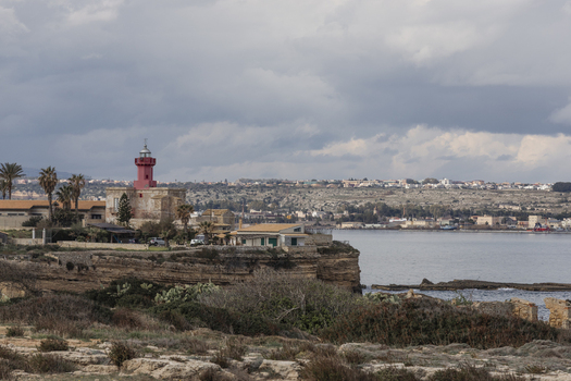 View of a red lighthouse on a rocky cliff overlooking the sea under a cloudy sky Syracuse Sicily Italy.