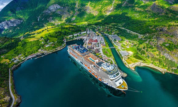 Aerial view of a large cruise ship docked in the deep blue waters of the Aurlandsfjord near the village of Flam surrounded by lush green mountains in Flam Vestland Norway. Aerial view of a large cruise ship docked in the deep blue waters of the Aurlandsfjord near the village of Flam surrounded by lush green mountains in Flam Vestland Norway.