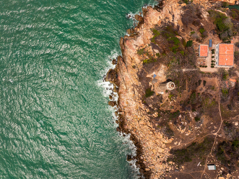 Aerial view of a lighthouse situated on a rugged rocky coastline with turquoise sea waves crashing against the cliffs and a small building with a red roof nearby in Vietnam. Aerial view of a lighthouse situated on a rugged rocky coastline with turquoise sea waves crashing against the cliffs and a small building with a red roof nearby in Vietnam.