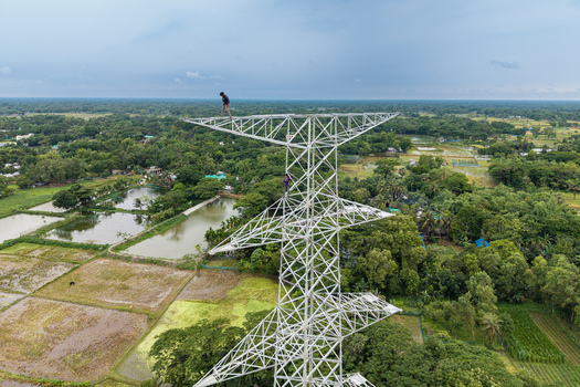 Trishal Upazila Bangladesh 13 May 2026 Aerial view of workers on a high voltage electricity pylon overlooking lush green fields and ponds under an overcast sky. Trishal Upazila Bangladesh 13 May 2026 Aerial view of workers on a high voltage electricity pylon overlooking lush green fields and ponds under an overcast sky.