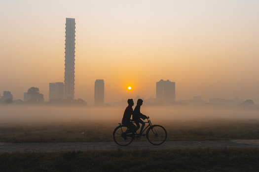 View of silhouettes of two people riding a bicycle through a misty landscape framed by modern skyscrapers under a glowing sunrise Kolkata West Bengal India. View of silhouettes of two people riding a bicycle through a misty landscape framed by modern skyscrapers under a glowing sunrise Kolkata West Bengal India.