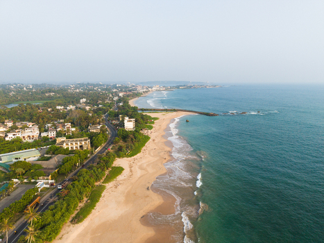 Aerial view of the sandy coastline with turquoise waves crashing on the shore and a coastal road lined with lush greenery and buildings in Galle Southern Province Sri Lanka.