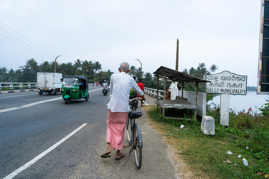 Galle Sri Lanka 29 January 2026 View of an elderly man with a bicycle on a road near the Galle Municipality sign with a green tuk tuk and palm trees.