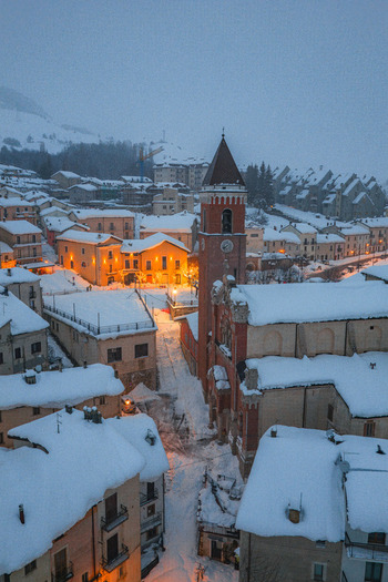 Aerial view of the snow covered town with its historic church tower illuminated at dusk surrounded by snow laden rooftops and mountain landscape in Rivisondoli Abruzzo Italy.