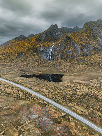 Aerial view of Lofoten landscape featuring a dramatic mountain with a waterfall a dark reflective lake and a winding road surrounded by autumn colored trees in Leknes Nordland Norway. Aerial view of Lofoten landscape featuring a dramatic mountain with a waterfall a dark reflective lake and a winding road surrounded by autumn colored trees in Leknes Nordland Norway.