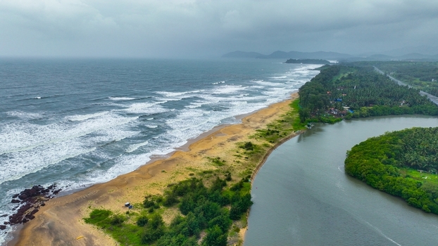 Aerial view of Galgibaga Beach where the river meets the sea featuring lush greenery sandy coastline and rolling waves under a cloudy sky in Canacona Goa India.