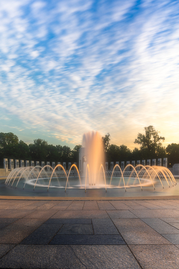 View of the World War II Memorial fountain with illuminated water jets and stone columns under a wispy sky at sunset in Washington District of Columbia United States. View of the World War II Memorial fountain with illuminated water jets and stone columns under a wispy sky at sunset in Washington District of Columbia United States.