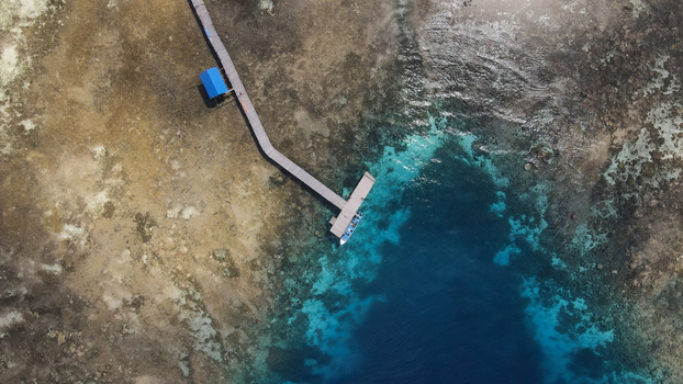 Aerial view of a wooden pier extending into the turquoise waters of Raja Ampat with a boat moored at the end surrounded by coral reefs in Raja Ampat Southwest Papua Indonesia.