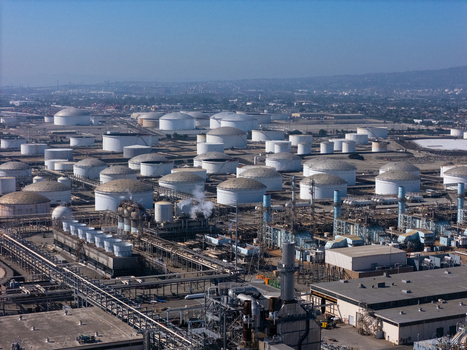 Aerial view of an industrial oil refinery featuring numerous white storage tanks complex piping systems and cooling towers under a clear sky in Carson California United States.