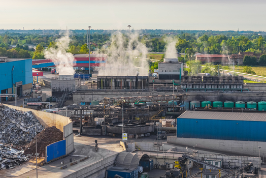 Aerial view of the Alfa Acciai steel plant featuring industrial cooling systems steam rising from production facilities and large scrap metal piles.