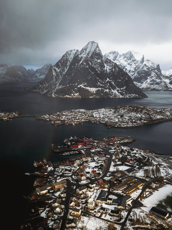 Aerial view of a small town nestled amongst snow dusted peaks and dark waters under a moody sky Reinebringen Nordland Norway.