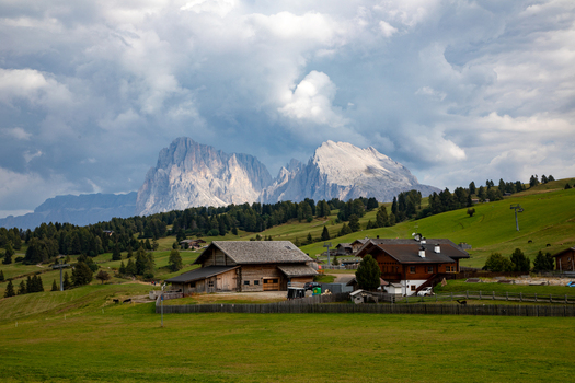 View of traditional wooden alpine houses in green meadows with the jagged peaks of the Dolomites under a dramatic cloudy sky in Rocca Pietore Veneto Italy. View of traditional wooden alpine houses in green meadows with the jagged peaks of the Dolomites under a dramatic cloudy sky in Rocca Pietore Veneto Italy.