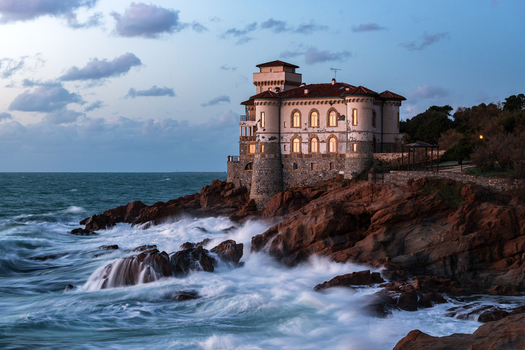 View of the historic Boccale Castle perched on the rocky cliffs with waves crashing against the shore at twilight in Calafuria Toscana Italy.