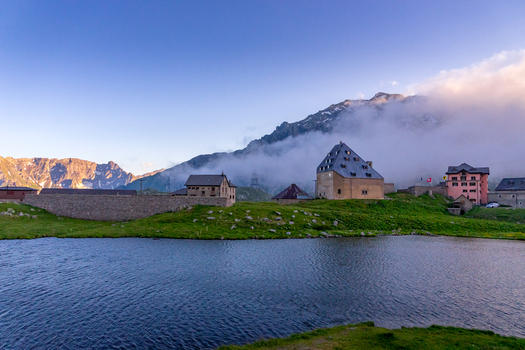 View of the historic Sankt Gotthard Pass buildings and a calm lake under a misty mountain peak in Airolo Ticino Switzerland.