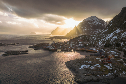 Aerial view of golden sunlight piercing through dramatic clouds over the Lofoten Islands casting a warm glow on the snow dusted mountains and the tranquil waters surrounding Tind Nordland Norway.