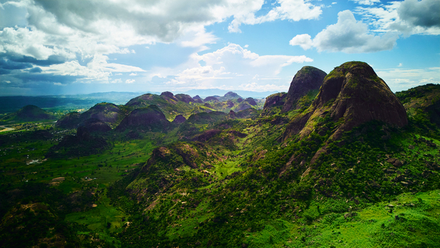 Aerial view of rugged verdant hills cascade under a vast sky dotted with fluffy clouds contrasting the rough terrain with soft heavenly light Abuja Nigeria.