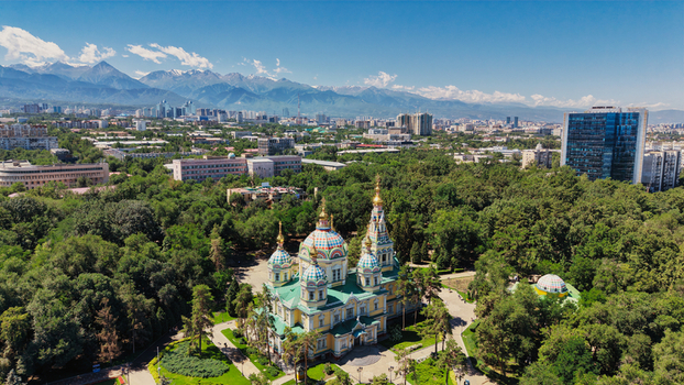 Aerial view of the colorful Zenkov Cathedral surrounded by lush green trees with the city skyline and mountain range in the background in Almaty Almaty Region Kazakhstan.
