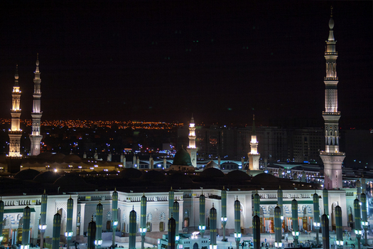 View of Al Masjid e Nabawi illuminated at night with its towering minarets and green dome glowing against the dark sky in Madinah Al Madinah Province Saudi Arabia.