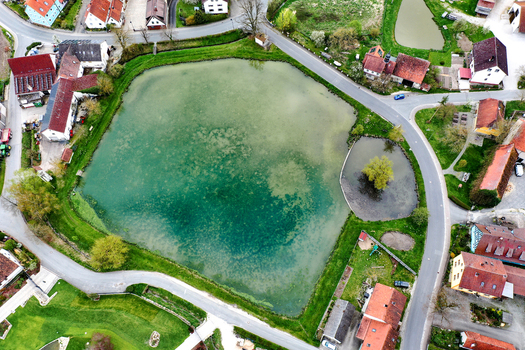 Aerial view of a green village pond surrounded by traditional houses with red roofs and winding roads in the rural landscape of Lonnerstadt Bayern Germany. Aerial view of a green village pond surrounded by traditional houses with red roofs and winding roads in the rural landscape of Lonnerstadt Bayern Germany.