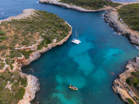 Aerial view of Cala Sa Nau with turquoise water rocky cliffs a white catamaran and a small boat in Felanitx Balearic Islands Spain. Aerial view of Cala Sa Nau with turquoise water rocky cliffs a white catamaran and a small boat in Felanitx Balearic Islands Spain.