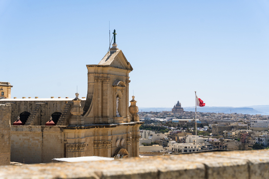 View of the historic Cathedral of the Assumption with its baroque facade and a Maltese flag waving against a clear blue sky Zebbug Gozo Region Malta. View of the historic Cathedral of the Assumption with its baroque facade and a Maltese flag waving against a clear blue sky Zebbug Gozo Region Malta.