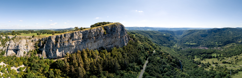 Aerial view of the massive limestone cliffs overlooking the lush Loue Valley with dense forests and a winding road under a clear blue sky in Mouthier Haute Pierre Bourgogne Franche Comte France. Aerial view of the massive limestone cliffs overlooking the lush Loue Valley with dense forests and a winding road under a clear blue sky in Mouthier Haute Pierre Bourgogne Franche Comte France.