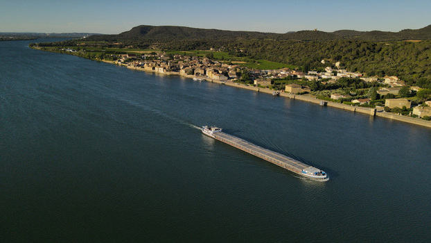 Aerial view of a cargo barge on the Rhone river passing the village with hills and vineyards in the background in Saint Etienne des Sorts Occitanie France. Aerial view of a cargo barge on the Rhone river passing the village with hills and vineyards in the background in Saint Etienne des Sorts Occitanie France.