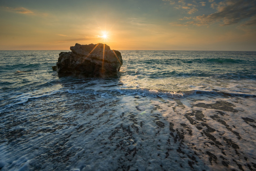 View of sunbeams bursting over a rock formation in the turquoise sea as waves foam against the dark shore Kathisma beach Kathisma Greece. View of sunbeams bursting over a rock formation in the turquoise sea as waves foam against the dark shore Kathisma beach Kathisma Greece.
