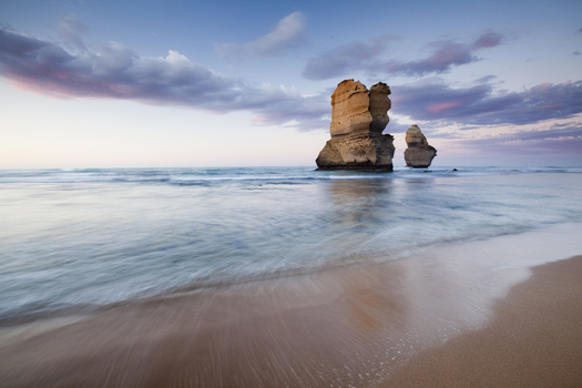 View of the limestone stacks of the Twelve Apostles Marine National Park rising from the ocean with a sandy beach and soft clouds at dusk Australia. View of the limestone stacks of the Twelve Apostles Marine National Park rising from the ocean with a sandy beach and soft clouds at dusk Australia.